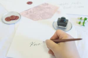 Eine Hand schreibt elegant mit einem Kalligrafiestift „Karo“ auf ein Stück Papier und erinnert damit an die feine Kunst der Hochzeitsplanung. Daneben sind ein Tintenfass, ein mit Wachs versiegelter Umschlag und ein Zweig kleiner Blumen sorgfältig auf einer makellos weißen Oberfläche arrangiert.
