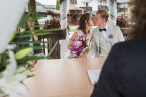 Ein Paar küsst sich während der Hochzeitszeremonie. Der Bräutigam trägt einen hellen Anzug und eine Fliege, während die Braut einen Strauß lila Blumen hält. Sie sitzen an einem Tisch unter einem wunderschön dekorierten Bogen, während im Hintergrund die feiernden Gäste zu sehen sind.