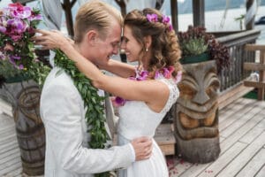 Braut und Bräutigam teilen einen zärtlichen Moment und berühren während ihrer Hochzeit ihre Stirn. Die Braut trägt einen Blumenkopfschmuck und eine Blumenkette, während der Bräutigam eine Blumenkette aus Grünzeug trägt. Sie stehen auf einem Holzdeck, das im Hintergrund mit Tiki-Schnitzereien geschmückt ist.