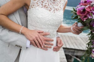 Ein Paar umarmt sich während seiner Hochzeit auf einem Holzsteg. Die Braut in einem weißen Spitzenkleid hält rosa Blumen in der Hand, während der Bräutigam einen hellgrauen Anzug trägt. Hinter ihnen bilden ein ruhiger See und majestätische Berge die perfekte Kulisse für diesen Moment der Liebe und Heirat.