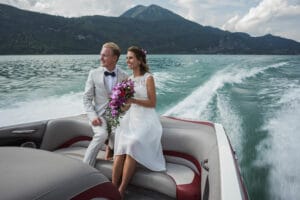 Eine Braut und ein Bräutigam sitzen nach ihrer Hochzeit strahlend vor Freude auf dem Heck eines Bootes. Die Braut, die einen Strauß lila Blumen umklammert, lächelt, während sie über einen See gleiten. Im Hintergrund erheben sich Berge unter einem teilweise bewölkten Himmel – eine perfekte Kulisse für dieses bedeutsame Heiraten-Abenteuer.