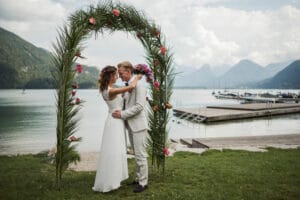 Braut und Bräutigam umarmen sich unter einem dekorativen Bogen mit Grünpflanzen und Blumen an einem ruhigen See mit Bergen im Hintergrund. In diesem wunderschönen Moment ihrer Hochzeit trägt der Bräutigam einen hellen Anzug und die Braut strahlt in ihrem weißen Kleid, während Paddleboards in der Nähe des Docks liegen.