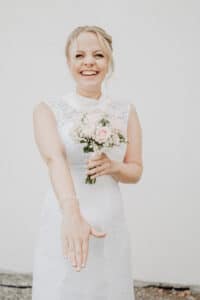 Eine lächelnde Braut in einem weißen Spitzenbrautkleid hält einen Strauß heller Blumen und streckt ihre Hand aus, um ihren Ring zu präsentieren. Sie fängt einen Moment der Freude ein, als sie sich vor einem schlichten weißen Hintergrund auf die wunderschöne Reise des Heiratens begibt.