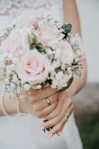 Eine Person in einem weißen Kleid hält einen Strauß hellrosa Rosen, gemischt mit weißem Schleierkraut, und verkörpert damit die Essenz romantischer Hochzeitsplanung. Ihre Hände, geschmückt mit einem schlichten Ring, umklammern den Strauß sanft vor einem sanft verschwommenen Hintergrund.