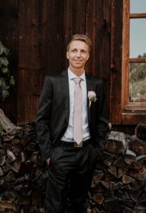 Ein Mann im Anzug mit hellrosa Krawatte und Boutonniere steht lächelnd da und verkörpert den Geist von Hochzeit, während er vor der Holzwand einer Hütte mit einem Stapel Brennholz unter einem Fenster posiert.
