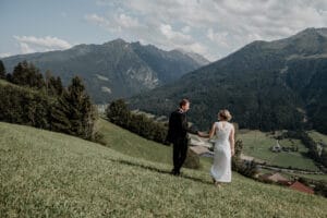 Ein Paar in eleganter Hochzeitskleidung geht an den Händen und anmutig einen grasbedeckten Hügel hinunter und verkörpert so seinen Traum vom Heiraten. Sie sind umgeben von einer malerischen Berglandschaft mit flauschigen Wolken und vereinzelten Bäumen und blicken auf ein kleines Dorf im Tal darunter.
