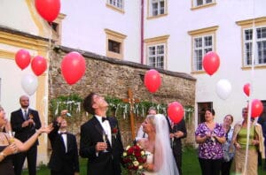 Braut und Bräutigam stehen lächelnd unter einem Himmel voller roter und weißer Luftballons, umgeben von Gästen in einem bezaubernden Innenhof. Sie feiern ihre Hochzeit, die Braut hält einen Blumenstrauß, während der Bräutigam einen dunklen Anzug trägt. Das Gebäude hinter ihnen hat hohe Fenster und eine Steinmauer.