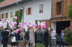 Eine Gruppe von Menschen in formeller Kleidung versammelt sich vor einem charmanten Holzgebäude und hält rosa und weiße Luftballons in den Händen. Die Stimmung ist feierlich, wahrscheinlich eine Hochzeit oder ein ähnliches Ereignis, bei dem die Gäste sich unterhalten und den Moment genießen.