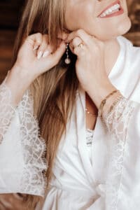 Eine lächelnde Frau rückt ihren Ohrring zurecht und verkörpert damit die Freude am Heiraten. Sie trägt ein weißes Gewand mit Spitzenärmeln und ein goldenes Armband. Ihr langes Haar fällt über ihre Schultern und ihre andere Hand zeigt einen Ehering und manikürte Nägel – eine perfekte Szene für eine tadellose Hochzeitsplanung.