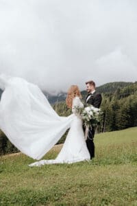 Mitten in der Hochzeitsplanung stehen Braut und Bräutigam liebevoll auf einer Wiese mit bewaldeten Bergen im Hintergrund. Das Kleid der Braut weht dramatisch im Wind, während sie einen großen Blumenstrauß hält. Beide lächeln unter einem bewölkten Himmel und genießen diesen perfekten Moment ihrer Hochzeit.