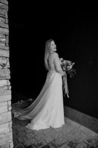 Eine Braut in einem eleganten Spitzenhochzeitskleid steht lächelnd mit dem Rücken zur Kamera und verkörpert die Freude einer Hochzeit. Sie hält einen Blumenstrauß und die Schleppe ihres Kleides fließt anmutig hinter ihr vor einer Steinmauer und einem dunklen Hintergrund.
