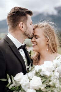 Ein Bräutigam im Smoking küsst die Stirn seiner lächelnden Braut, die einen Strauß weißer Rosen in der Hand hält. Sie sind draußen mit nebligen Bergen im Hintergrund, was eine romantische und ruhige Kulisse schafft – ein perfekter Moment auf ihrer bezaubernden Hochzeitsreise.