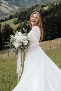 Eine Braut in einem Spitzenhochzeitskleid hält einen Strauß weißer Blumen und lächelt freudig. Sie steht auf einer Wiese mit sanften Hügeln und Bäumen und fängt die Essenz einer Hochzeit in dieser malerischen, natürlichen Umgebung ein.