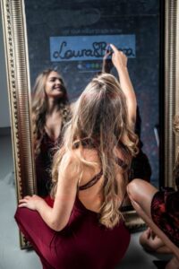 Eine Frau in einem roten Kleid kniet vor einem großen, verzierten Spiegel und lächelt ihr Spiegelbild an. Sie malt „Laura & Rosa“ auf das Glas, ihr langes Haar fällt ihr über die Schulter – ein Moment der Freude inmitten des Trubels der Hochzeitsplanung.