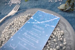 Eine Hochzeitsmenükarte liegt auf einem dekorativen Teller, der mit kleinen weißen Beeren geschmückt ist, umrahmt von einer Gabel auf einer strukturierten blauen Tischdecke. Das Menü zeigt elegante Grafiken im Winterthema mit Schneeflocken und verleiht der Hochzeitsplanung einen Hauch von Magie.