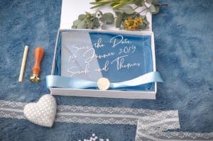 Eine „Save the Date“-Karte für Sarahs und Thomas‘ Hochzeit am 19. Januar 2019. Die Karte, perfekt für alle, die gerade mit der Hochzeitsplanung beschäftigt sind, ist mit einer blauen Schleife und einem Wachssiegel verziert. Sie liegt in einer Schachtel auf einer hellblauen Oberfläche, umgeben von eleganten Dekorationsartikeln.