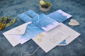 Eine Hochzeitseinladung auf blauem Stoffhintergrund weckt den Zauber eines Winterthemas mit Bäumen und Schneeflocken. Sie feiert die Liebe und enthält einen Umschlag mit Kalligraphie, zwei Goldmünzen, eine Perlenkette und eine herzförmige Dekoration – perfekt für jedes Paar, das seine Traumhochzeit plant.