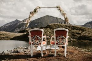 Zwei elegante Stühle mit Blick auf eine malerische Berglandschaft an einem See, perfekt für eine Hochzeit. Die Stühle sind mit Blumenkränzen und Schildern mit der Aufschrift „Bräutigam“ und „Braut“ dekoriert. Ein dekorativer Bogen mit Blumen vervollständigt die intime Atmosphäre im Freien, ideal für diejenigen, die heiraten möchten.