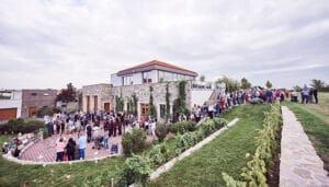 Eine große Menschenmenge versammelt sich auf einem Weg und einer Rasenfläche vor einem modernen zweistöckigen Steingebäude mit rotem Ziegeldach, ideal für eine Hochzeit. Die Szene entfaltet sich unter einem bewölkten Himmel, mit üppigem Grün und gepflegten Landschaftsgestaltungen, die die malerische Kulisse umgeben.