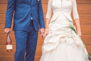 Ein Paar in formeller Kleidung steht Händchen haltend da und ist bereit, seine Hochzeit zu feiern. Der Mann im blauen Anzug hält eine Flasche Roséwein, während die Frau ein weißes Hochzeitskleid mit Blumendetails trägt und einen Strauß grasähnlicher Pflanzen trägt. Ein hölzerner Hintergrund vervollständigt die Szene.