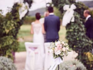 Ein Paar steht während seiner Hochzeit vor einem mit Grünpflanzen und Bändern geschmückten Bogen. Im Vordergrund ist ein Blumenarrangement mit weißen und rötlichen Blumen zu sehen, das die Essenz der sorgfältigen Hochzeitsplanung einfängt, die ihre besondere Zeremonie im Freien unvergesslich machte.