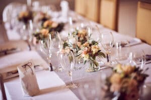 Ein langer, elegant gedeckter Esstisch für eine formelle Hochzeit bietet ordentlich angeordnete Weingläser, gefaltete Servietten und kleine Blumengestecke mit zarten Pfirsich- und Cremeblüten, alles auf einer weißen Tischdecke mit grauem Läufer. Diese exquisite Kulisse ist perfekt für alle, die sich akribisch auf die Hochzeitsplanung konzentrieren.