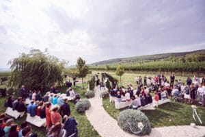 Eine große Hochzeitszeremonie im Freien findet in einem Weinberg statt und zeigt eine sorgfältige Hochzeitsplanung. Die Gäste sitzen auf weißen Bänken entlang eines geschwungenen Weges, umgeben von Grün. Das Paar steht unter einem bewölkten Himmel an einem Altar. Hügel und Weinreben dienen als romantische Kulisse für diese Hochzeit.