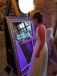 Eine Frau in einem weißen Kleid berührt einen großen interaktiven Spiegel mit violetten Lichtern und unterschreibt bei einer Hochzeit mit ihrem Finger ihren Namen. Ein junges Mädchen steht daneben und beobachtet. Die Kulisse besteht aus Backsteinmauern und Holzelementen und suggeriert einen rustikalen Ort, der perfekt zum Heiraten ist.
