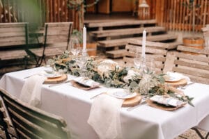 Ein wunderschön gedeckter Esstisch im Freien mit Holzstühlen, ideal für eine Hochzeit, mit weißen Kerzenleuchtern, Grünpflanzen, Blumen und elegantem Geschirr mit gefalteten Servietten auf Tellern. Die Umgebung strahlt mit Holzstufen und Laternen im Hintergrund rustikalen Charme aus.