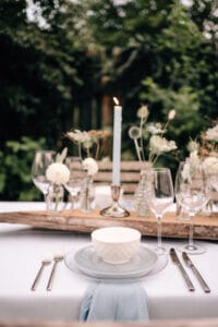Ein schön gedeckter Esstisch im Freien verströmt den Charme einer Hochzeit. Er verfügt über eine brennende Kerze, weiße Blumen in Vasen, Weingläser und eine weiße Schüssel auf gestapelten Tellern mit Besteck. Tischdecke und Serviette sind hellblau, perfekt für jede Hochzeitsplanung inmitten üppigen Grüns.