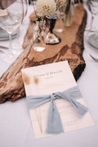 Eine bezaubernde Hochzeitseinladung für „Adriana & Benedikt“, die für den 14. Juni 2024 angesetzt ist, ruht elegant auf einem rustikalen Holztisch. Mit kleinen Blumenvasen und einem blauen Band, das mit dekorativen getrockneten Gräsern verflochten ist, ist sie perfekt für diejenigen, die sich tief mit den Nuancen der Hochzeitsplanung auskennen.