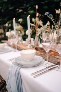 Ein wunderschön gedeckter Tisch im Freien mit weißem und hellblauem Geschirr, Silberbesteck, Kristallgläsern und blauen Kerzen schafft die perfekte Kulisse für eine Hochzeit. Trockenblumen in Vasen zieren das rustikale Holzgesteck auf einer weißen Tischdecke und machen es zum idealen Ort für jede Hochzeitsfeier.
