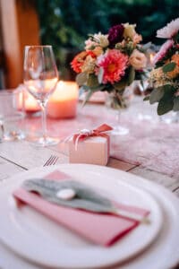Ein wunderschön gedeckter Esstisch mit einem weißen Teller und einer rosa Serviette, geschmückt mit Grünpflanzen. Daneben steht eine kleine Geschenkbox mit einer Schleife. Auf dem Tisch stehen elegante Gläser, Kerzen und farbenfrohe Blumenarrangements – perfekt für eine romantische Hochzeitsfeier.