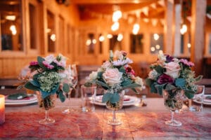 Ein für eine festliche Hochzeit gedeckter Tisch besteht aus drei eleganten Blumenarrangements in Glasvasen. Die Arrangements umfassen Rosen und Grünpflanzen. Warme Beleuchtung und Holzdekor schaffen im Hintergrund eine gemütliche Atmosphäre, perfekt für alle, die Inspiration bei der Hochzeitsplanung suchen.