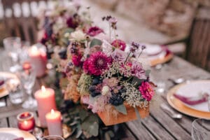 Ein rustikaler Holztisch ist mit einem farbenfrohen Blumengesteck aus rosa, violetten und weißen Blumen geschmückt, akzentuiert durch Grünpflanzen und Beeren. Perfekt für eine Hochzeit, umfasst das Ambiente brennende Kerzen und elegantes Geschirr und schafft eine warme und einladende Atmosphäre, die für Heiratsfeiern geeignet ist.