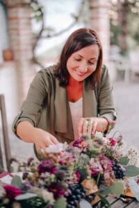 Eine Frau mit braunem Haar und einer grünen Jacke lächelt, während sie im Freien auf einem Tisch ein farbenfrohes Blumenarrangement mit violetten Blumen und Grünpflanzen arrangiert, perfekt für eine Hochzeit.