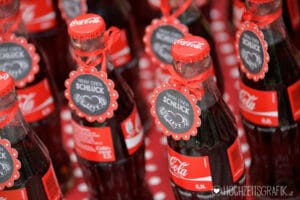 Nahaufnahme mehrerer Coca-Cola-Flaschen mit roten Verschlüssen und dekorativen Etiketten auf rotem gepunktetem Stoff, perfekt für eine Hochzeit. Die Etiketten haben ein Design im Tafelstil mit deutschem Text und verleihen jeder Hochzeitsplanung eine charmante Note.