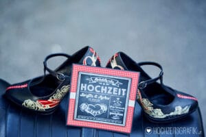 Ein Paar schwarze High Heels mit Blumenmustern wird um eine quadratische rot-weiße Einladung zu einer Hochzeit herum ausgestellt. Die Einladung ist im Tafel-Stil gestaltet und hat einen handgeschriebenen Text in Deutsch, perfekt für Ihre Hochzeitsfeier.