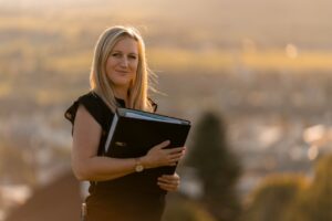 Eine Frau mit langen blonden Haaren steht im Freien und hält einen Ordner, möglicherweise mit Hochzeitsplänen, vor einem verschwommenen Hintergrund aus Bäumen und einer fernen Landschaft. Sie trägt ein schwarzes ärmelloses Oberteil und lächelt sanft im warmen Licht.