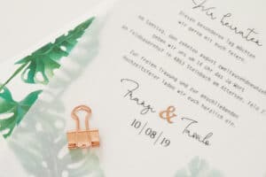 Eine Hochzeitseinladung mit einer durchscheinenden Überlagerung mit grünen Blattmotiven, perfekt für Ihre Hochzeit. Sie enthält deutschen Text und ein Datum vom 10.08.19. In der oberen linken Ecke ist eine kleine kupferfarbene Heftklammer angebracht, die Ihrer Hochzeitsplanung eine schicke Note verleiht.