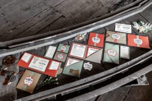 Eine Sammlung farbenfroher Hochzeitseinladungen und Umschläge ist kunstvoll auf einer rustikalen Holzoberfläche arrangiert und spiegelt perfekt die Essenz der Hochzeitsplanung wider. Verschiedene Designs und Motive leuchten hell, und einige Umschläge öffnen sich und geben Karten frei, die zwischen kleinen Blumendekorationen eingebettet sind.
