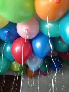 Eine bunte Auswahl an Heliumballons, perfekt für eine Hochzeitsfeier, schwebt an der Decke. In den Farbtönen Rot, Blau, Grün, Gelb und Rosa ist jeder Ballon elegant an einer herabhängenden Schnur befestigt.