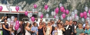 Eine Hochzeitsfeier findet im Freien vor einer Bergkulisse statt. Die Gäste halten rosa und weiße Luftballons, während Braut und Bräutigam lächelnd nebeneinander stehen. Im Hintergrund liegt ein Boot vor Anker, was die sonnige, festliche Szene noch verstärkt.