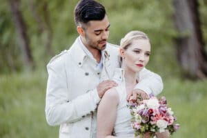Ein Paar steht auf einer Wiese und verkörpert die Essenz von Hochzeit. Der Mann, der eine weiße strukturierte Jacke trägt, umarmt die Frau sanft von hinten. Sie hält einen Strauß rosa und weißer Blumen und trägt ein weißes Kleid. Beide haben einen heiteren Gesichtsausdruck, im Hintergrund sind die Bäume verschwommen.