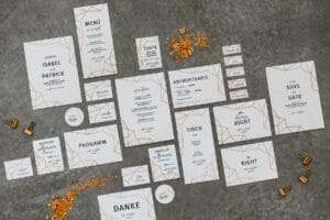 Eine Sammlung elegant gestalteter Hochzeitspapeterieartikel, perfekt für die Hochzeitsplanung, darunter Einladungen, Menükarten, Tischnummern und Save-the-Date-Karten. Sie sind auf einer grauen Oberfläche angeordnet und überall sind goldene dekorative Elemente verstreut.