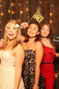 Drei Frauen posieren fröhlich in einer Fotokabine bei einer Hochzeit, umgeben von Requisiten. Eine hält eine originelle Brille, eine andere ein „Team Braut“-Schild und die dritte eine Sprechblase mit der Aufschrift „Ich bin die Nächste“. Sie stehen vor einem Hintergrund, der mit funkelnden Lichterketten geschmückt ist und die Freude der Hochzeit einfängt.