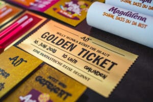 Nahaufnahme eines „Goldenen Tickets“ für eine Hochzeitsveranstaltung am 16. Juni 2018 um 16 Uhr in Gmunden. Auf dem Ticket steht „Willy Wonka küsst die Braut“, umgeben von bunten Karten und Stiften, die die skurrile Essenz einzigartiger Hochzeitsplanung einfangen.