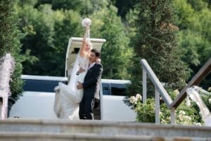 Braut und Bräutigam feiern freudig ihre Hochzeit vor einer weißen Limousine. Die Braut reckt ihren Brautstrauß in die Luft, während der Bräutigam sie anlächelt und stützt. Sie sind umgeben von üppigem Grün und Treppen im Vordergrund.