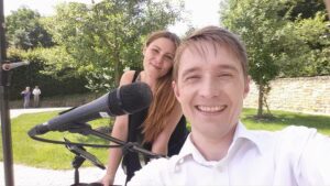 Ein lächelnder Mann und eine lächelnde Frau machen an einem sonnigen Tag im Freien ein Selfie und halten so ihren besonderen Hochzeitsmoment fest. Der Mann steht im Vordergrund und hält die Kamera. Neben der Frau ist ein Mikrofon zu sehen, was darauf hindeutet, dass sie ihre Freude mit ihren Lieben teilen. Sie stehen in einem Garten mit üppigen grünen Bäumen und einer Steinmauer im Hintergrund.