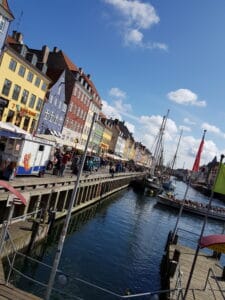 Ein malerischer Blick auf eine farbenfrohe Uferpromenade mit Reihen lebhafter Gebäude entlang eines Kanals bietet die perfekte Kulisse für eine Traumhochzeit. Ein großes Schiff liegt daneben und Menschen schlendern unter einem klaren blauen Himmel über die Holzpromenade. Flaggen wehen anmutig am Wasser und verleihen jeder Hochzeitsplanung Charme.